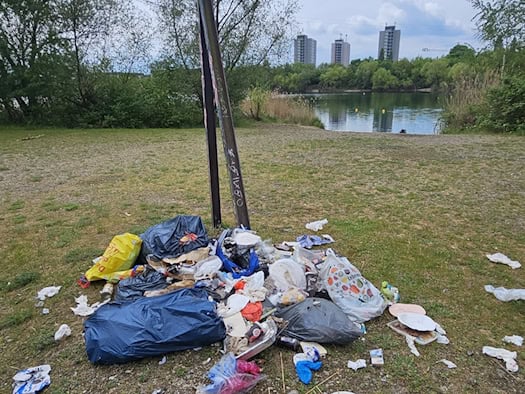 Ein großer Haufen wilder Müll aus Plastiktüten, Papptellern und Dosen auf einer Wiese. Im Hintergrund sieht man einen See, grüne Bäume und drei weit entfernte Wohnhochhäuser unter bewölktem Himmel.
