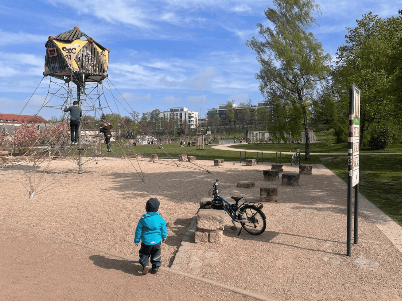 Eine Außenaufnahme eines weitläufigen Spielplatzes an einem leicht bewölkten Frühlingstag. Im linken Bereich steht ein großes Kletter-Seilnetz, das pyramidenförmig zu einem hölzernen Spielhaus auf Stelzen hochführt. Im Vordergrund läuft ein kleines Kind in einer hellblauen Jacke und dunkler Hose.