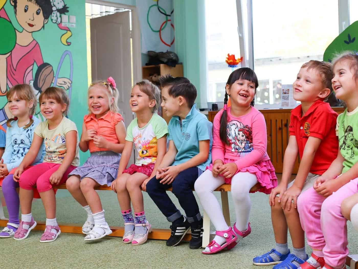 Eine Gruppe von acht fröhlichen Jungen und Mädchen sitzt nebeneinander auf einer Holzbank in einem bunten Kindergartenraum und lacht. Im Hintergrund ist eine farbenfroh bemalte Wand zu sehen.