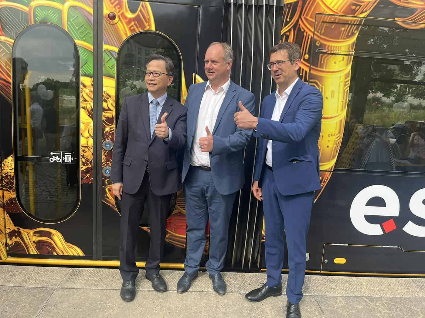 Three men in elegant suits stand smiling side by side in front of a black-and-yellow-wrapped tram and give a thumbs-up together.