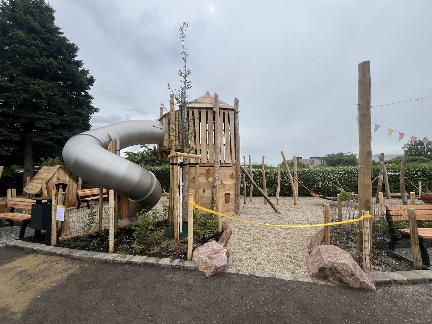 Ein großer Spielplatz-Kletterturm aus hellem Naturholz mit einer silbernen, spiralförmigen Röhrenrutsche. Ein gelbes Absperrband trennt den Bereich im Vordergrund ab.