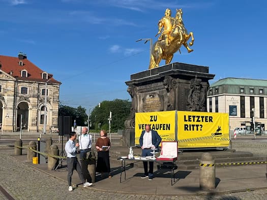 Das vergoldete Reiterstandbild des Goldenen Reiters in Dresden ist mit einem großen gelben Banner mit der Aufschrift „ZU VERKAUFEN. LETZTER RITT?“ verhüllt. Davor stehen Personen an einem Tisch mit Tafelsilber.