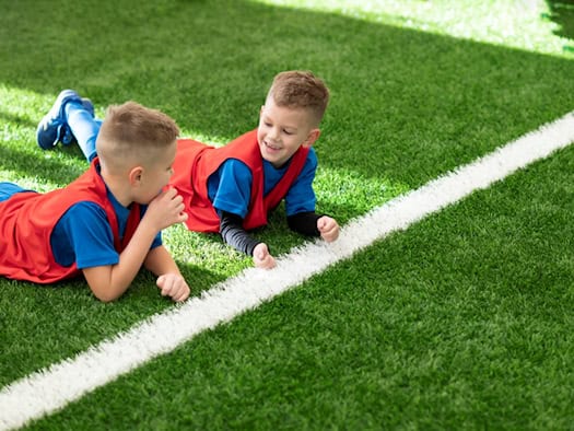 Zwei kleine Jungen in blauen Trikots und roten Trainingswesten liegen lächelnd nebeneinander auf einem saftig grünen Kunstrasenplatz direkt neben einer breiten, weißen Spielfeldlinie.