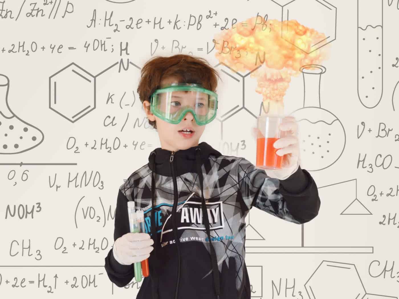 Ein Junge mit grüner Schutzbrille und Handschuhen hält Reagenzgläser und ein Becherglas mit orangefarbener Flüssigkeit, aus der eine kleine Explosionswolke aufsteigt. Der helle Hintergrund ist voller mathematischer und chemischer Formeln.