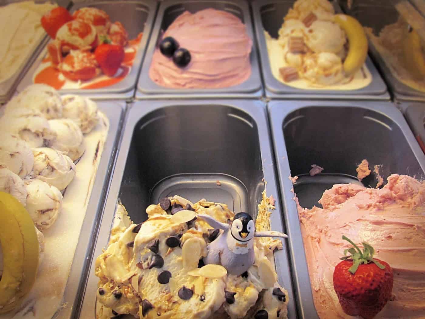 Eine bunte Auswahl verschiedener Speiseeis-Sorten in den Edelstahlbehältern einer Eistheke. Zu sehen sind unter anderem Erdbeereis mit einer echten Frucht, helles Eis mit Bananen- und Schokodekor sowie eine kleine Pinguin-Plastikfigur als Dekoration auf einer cremigen Eissorte.