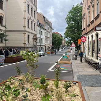 Blick entlang der Louisenstraße in Dresden. Auf der rechten Straßenseite, wo früher Autos parkten, stehen nun moderne, rostbraune Pflanzkübel mit jungen Grünpflanzen sowie Sitzgelegenheiten vor den typischen Neustadt-Altbauten.