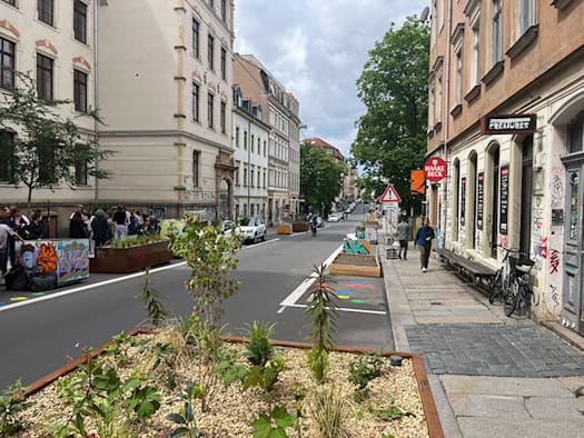 Blick entlang der Louisenstraße in Dresden. Auf der rechten Straßenseite, wo früher Autos parkten, stehen nun moderne, rostbraune Pflanzkübel mit jungen Grünpflanzen sowie Sitzgelegenheiten vor den typischen Neustadt-Altbauten.