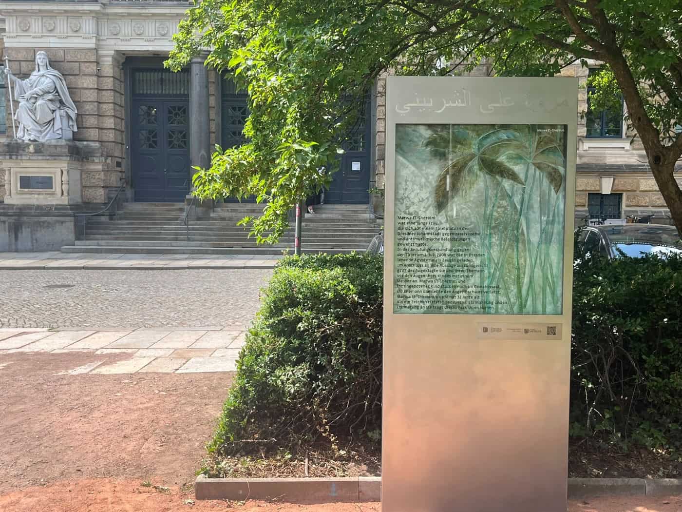 Eine moderne Gedenkstele aus Metall und Glas steht im Marwa-El-Sherbini-Park. Auf dem Glas sind grüne Pflanzenmotive und ein biografischer Text zu sehen. Im Hintergrund ist der steinerne Eingang des Landgerichts Dresden erkennbar.