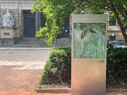 Eine moderne Gedenkstele aus Metall und Glas steht im Marwa-El-Sherbini-Park. Auf dem Glas sind grüne Pflanzenmotive und ein biografischer Text zu sehen. Im Hintergrund ist der steinerne Eingang des Landgerichts Dresden erkennbar.