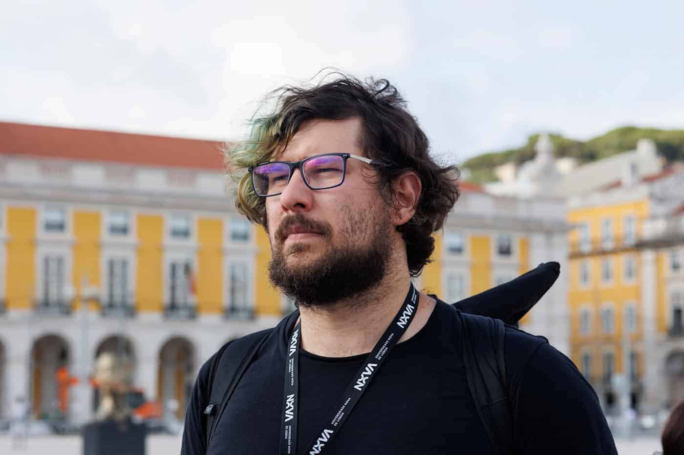 Mathias Fröck, stellvertretender Verwaltungsratsvorsitzender des Studentenwerks Dresden Ein Porträt eines Mannes mit dunklem, welligem Haar, Brille und einem Vollbart, der im Profil nach links blickt. Er trägt ein schwarzes T-Shirt und ein bedrucktes Schlüsselband um den Hals. Im unscharfen Hintergrund ist ein großer öffentlicher Platz mit gelben, historischen Gebäuden und Rundbögen zu sehen.