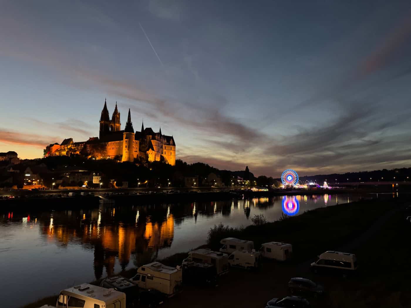 Foto: beleuchtete Burg von Meißen, thront über dem Fluss, in dem sich die Lichter der Burg, der Stadt und dem bunten Riesenrad spiegeln. Der Himmel ist abendlich gefärbt und fast dunkel.
