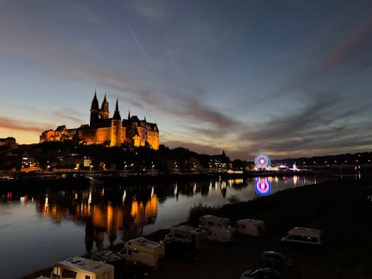 Foto: beleuchtete Burg von Meißen, thront über dem Fluss, in dem sich die Lichter der Burg, der Stadt und dem bunten Riesenrad spiegeln. Der Himmel ist abendlich gefärbt und fast dunkel.