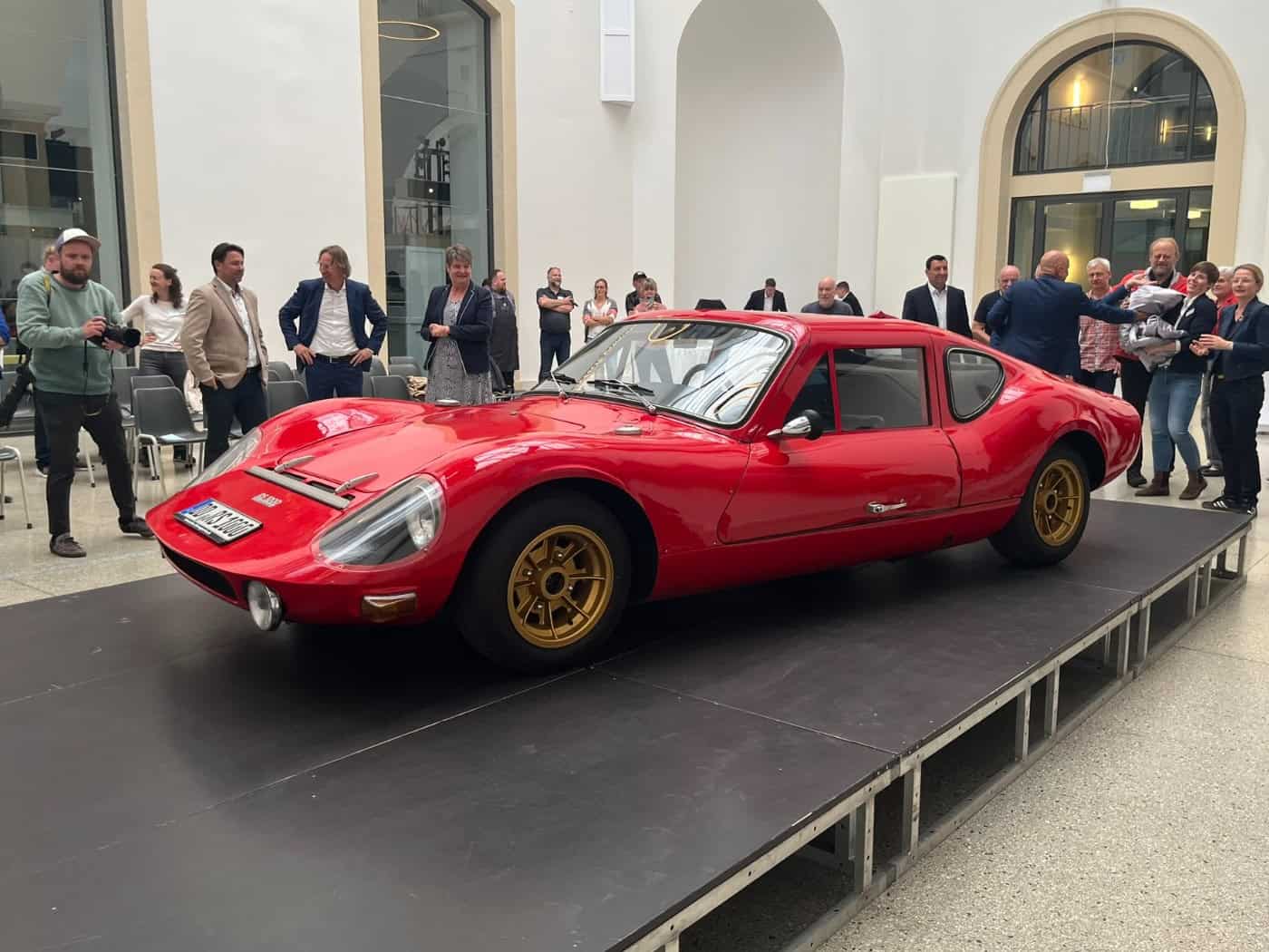 Ein flacher, knallroter DDR-Sportwagen vom Typ Melkus RS 1000 steht im Rampenlicht auf einer niedrigen, schwarzen Bühne. Im Hintergrund stehen in einem hellen, weitläufigen Lichthof des Verkehrsmuseums Dresden zahlreiche Zuschauer, Fotografen und Museumsmitarbeiter im Halbkreis um das Fahrzeug.