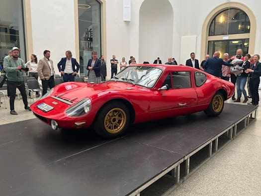 Ein flacher, knallroter DDR-Sportwagen vom Typ Melkus RS 1000 steht im Rampenlicht auf einer niedrigen, schwarzen Bühne. Im Hintergrund stehen in einem hellen, weitläufigen Lichthof des Verkehrsmuseums Dresden zahlreiche Zuschauer, Fotografen und Museumsmitarbeiter im Halbkreis um das Fahrzeug.