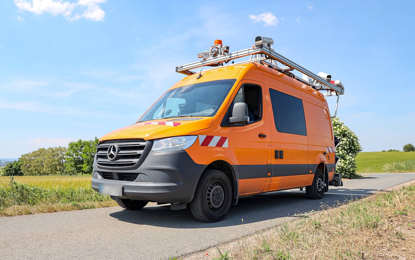 Ein orangefarbener Mercedes-Transporter mit rot-weißer Warnmarkierung und einer großen Metallkonstruktion inklusive Kameras und Sensoren auf dem Dach steht auf einer asphaltierten Straße vor Feldern unter blauem Himmel.