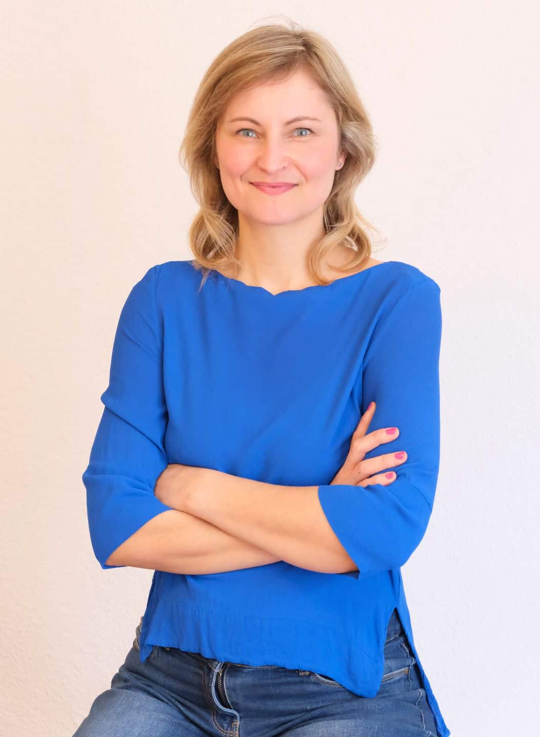 Porträtfoto einer lächelnden Frau in Jeans und blauem Oberteil, die Arme vorm Körper verschränkt