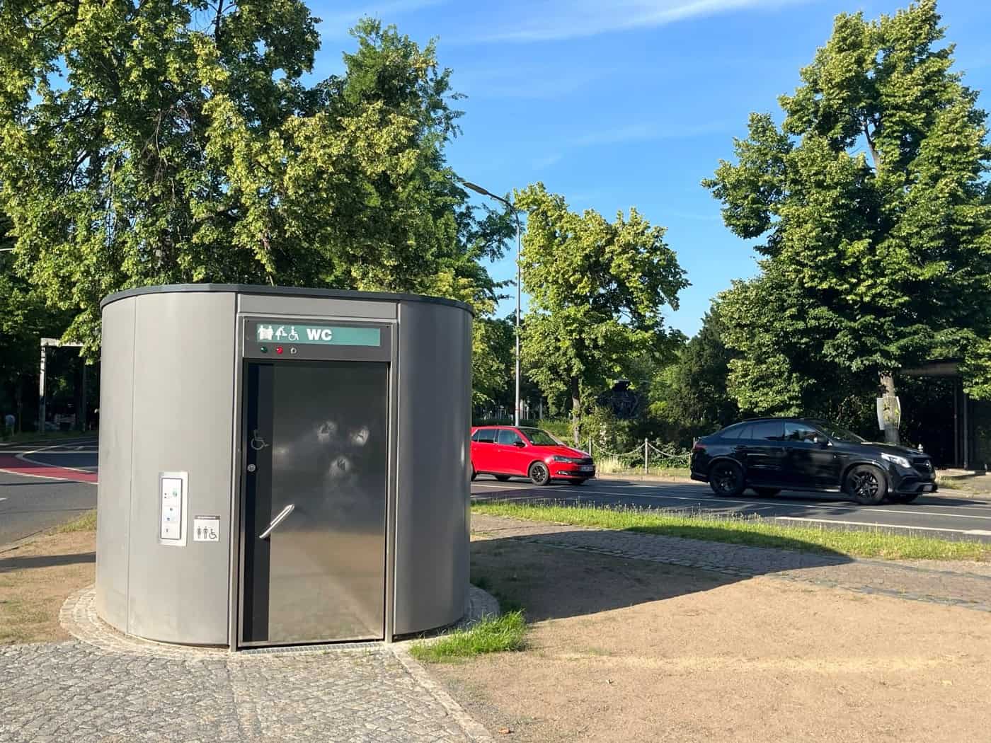Eine moderne, ovale Unisex-Toilette aus dunkelgrauem Sichtbeton mit einer Edelstahltür steht auf einer gepflasterten Verkehrsinsel. Über der Tür ist ein grünes Schild mit „WC“ und Piktogrammen. Im Hintergrund fahren Autos an Bäumen vorbei.
