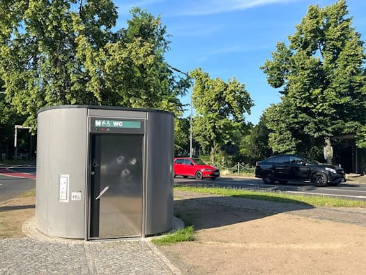Eine moderne, ovale Unisex-Toilette aus dunkelgrauem Sichtbeton mit einer Edelstahltür steht auf einer gepflasterten Verkehrsinsel. Über der Tür ist ein grünes Schild mit „WC“ und Piktogrammen. Im Hintergrund fahren Autos an Bäumen vorbei.