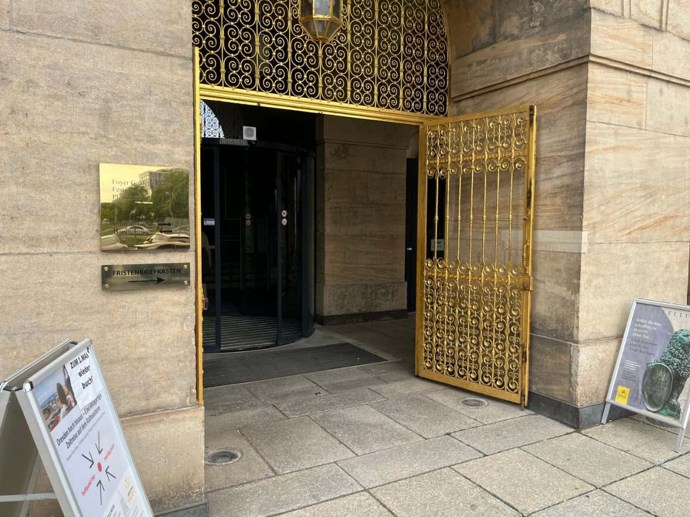 Ein historisches Sandsteinportal mit einem geöffneten, kunstvoll verzierten goldenen Metalltor und einer Drehtür. Links an der Wand hängt ein goldenes Schild mit der Aufschrift „Foyer Goldene Pforte“ und „Plenarsaal“.