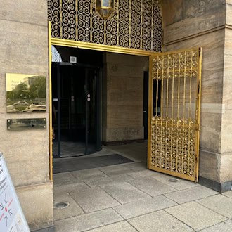 Ein historisches Sandsteinportal mit einem geöffneten, kunstvoll verzierten goldenen Metalltor und einer Drehtür. Links an der Wand hängt ein goldenes Schild mit der Aufschrift „Foyer Goldene Pforte“ und „Plenarsaal“.