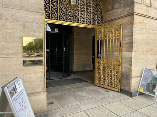 Ein historisches Sandsteinportal mit einem geöffneten, kunstvoll verzierten goldenen Metalltor und einer Drehtür. Links an der Wand hängt ein goldenes Schild mit der Aufschrift „Foyer Goldene Pforte“ und „Plenarsaal“.