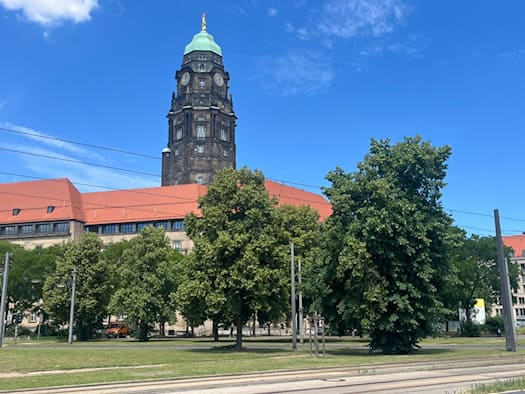 Der dunkle Steinturm des Neuen Rathauses in Dresden ragt hinter großen, grünen Laubbäumen in einen blauen Sommerhimmel. Auf der Turmspitze befindet sich eine hellgrüne Kuppel. Im Vordergrund verlaufen Straßenbahnschienen.