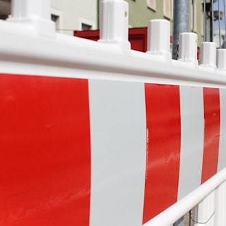 Nahaufnahme einer rot-weiß gestreiften Baustellenabsperrung aus Kunststoff. Im verschwommenen Hintergrund sind ein grauer Bauzaun, ein roter Container und die Fassade eines Wohngebäudes zu sehen.