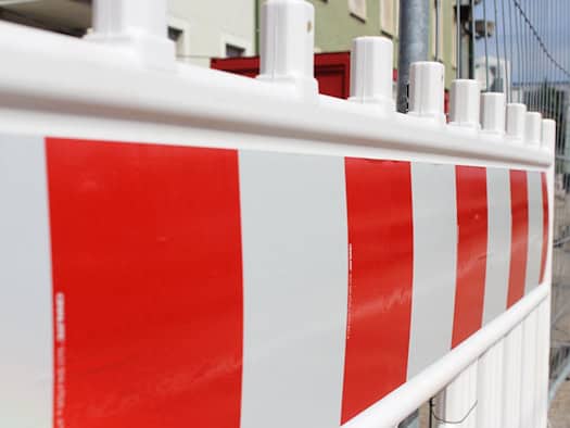 Nahaufnahme einer rot-weiß gestreiften Baustellenabsperrung aus Kunststoff. Im verschwommenen Hintergrund sind ein grauer Bauzaun, ein roter Container und die Fassade eines Wohngebäudes zu sehen.