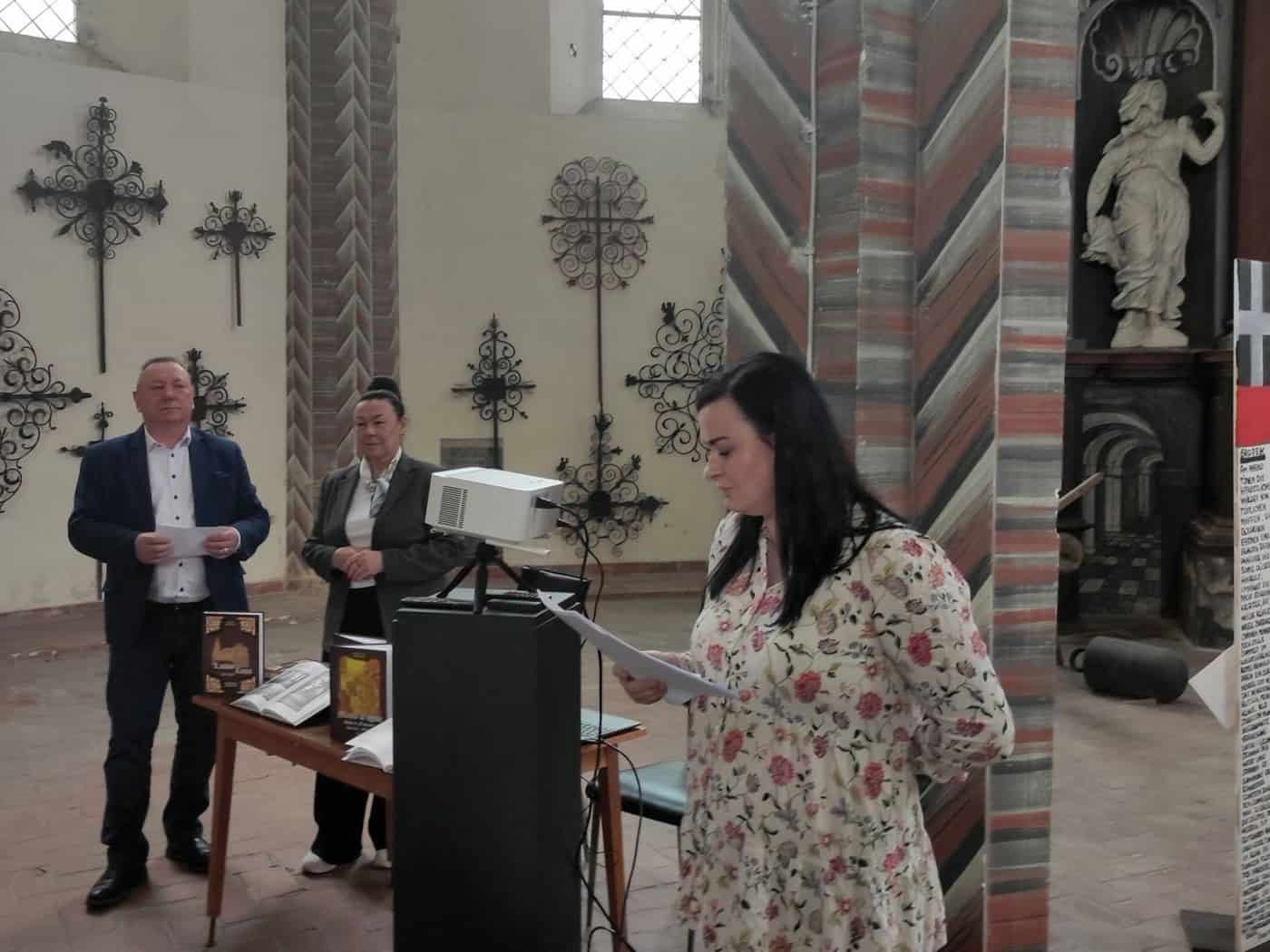 Eine Frau schaut auf Papierblätter in ihrer Hand, in einem Kirchenraum, im Hintergrund ein Beamer, Büchertisch und 2 weitere Personen