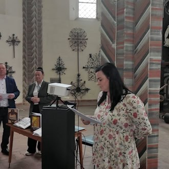 Eine Frau schaut auf Papierblätter in ihrer Hand, in einem Kirchenraum, im Hintergrund ein Beamer, Büchertisch und 2 weitere Personen