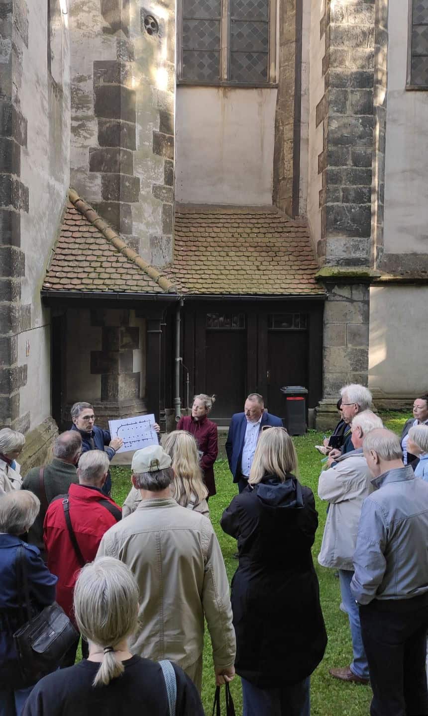 eine Gruppe steht vor einem Seitenbereich einer Kirche, vor der Gruppe steht ein Mann und zeigt einen Kirchengrundriss