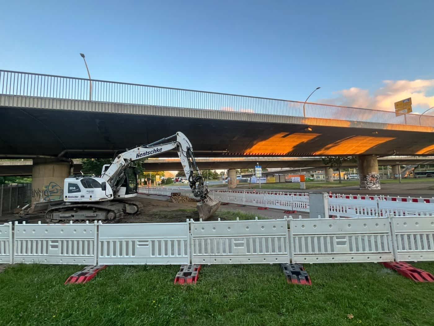 Ein weißer Kettenbagger steht unter der Brücke Budapester Straße in Dresden; der Baustellenbereich ist im Vordergrund mit weißen Kunststoff-Absperrgittern gesichert.