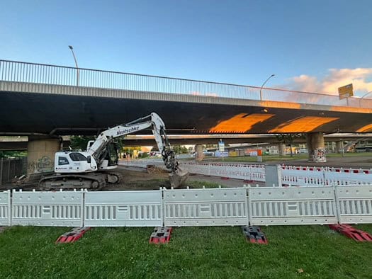 Ein weißer Kettenbagger steht unter der Brücke Budapester Straße in Dresden; der Baustellenbereich ist im Vordergrund mit weißen Kunststoff-Absperrgittern gesichert.
