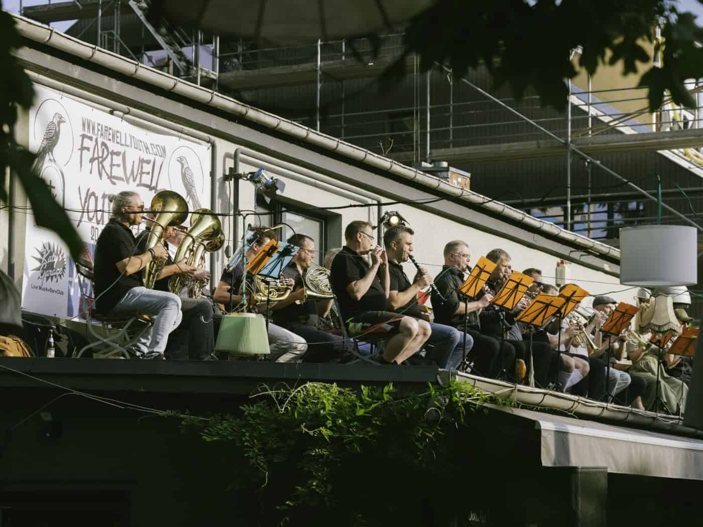 Mehrere Musiker stehen mit ihren Blechblasinstrumenten auf dem flachen Dach des Gastronomiebetriebs „Katy's Garage“ im Freien. Im Hintergrund sind die bunten Fassaden und Fenster der umliegenden Wohnhäuser in der Dresdner Neustadt sowie grünes Laub von Bäumen zu sehen.