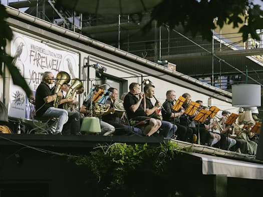 Mehrere Musiker stehen mit ihren Blechblasinstrumenten auf dem flachen Dach des Gastronomiebetriebs „Katy's Garage“ im Freien. Im Hintergrund sind die bunten Fassaden und Fenster der umliegenden Wohnhäuser in der Dresdner Neustadt sowie grünes Laub von Bäumen zu sehen.