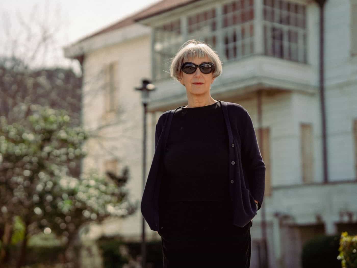 Ein Porträt von Olga Martynova mit kurzen, grauen Haaren und einer dunklen Sonnenbrille. Sie trägt eine schwarze Bluse und eine dunkle Strickjacke und blickt direkt in die Kamera. Im unscharfen Hintergrund ist ein helles, historisches Gebäude mit einem großen Holzbalkon zu sehen.