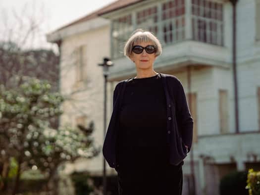 Ein Porträt von Olga Martynova mit kurzen, grauen Haaren und einer dunklen Sonnenbrille. Sie trägt eine schwarze Bluse und eine dunkle Strickjacke und blickt direkt in die Kamera. Im unscharfen Hintergrund ist ein helles, historisches Gebäude mit einem großen Holzbalkon zu sehen.
