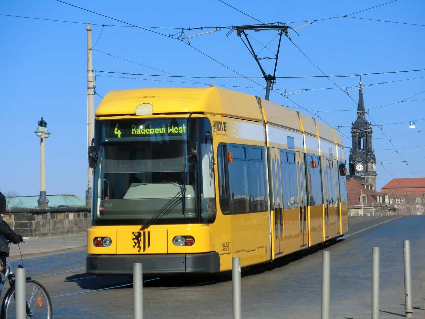 Eine gelbe Straßenbahn der Dresdner Verkehrsbetriebe (DVB) auf der Linie 4 Richtung Radebeul West fährt bei blauem Himmel über eine Brücke in Dresden, im Hintergrund ist die Dreikönigskirche zu sehen.
