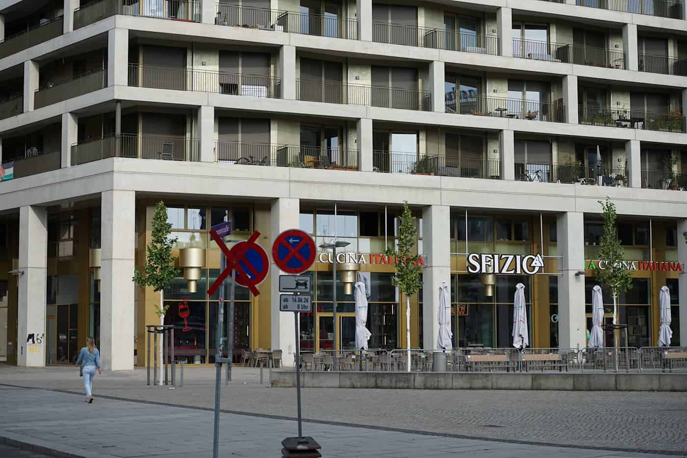 Blick auf einen gepflasterten Platz mit Verkehrsschildern im Vordergrund. Im Hintergrund befindet sich ein großes Wohn- und Geschäftshaus mit Außenplätzen des Restaurants „Sfizio“. Eine Passantin läuft von links durch das Bild.