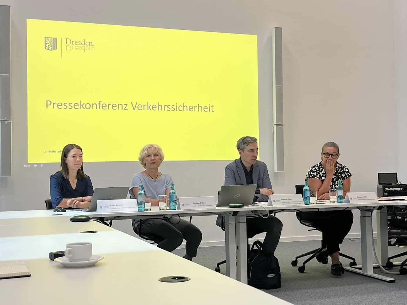 Vier Personen sitzen an einem langen weißen Tisch vor einer gelben Projektionswand mit der Aufschrift „Pressekonferenz Verkehrssicherheit“. Auf dem Tisch stehen Laptops und Wasserflaschen.