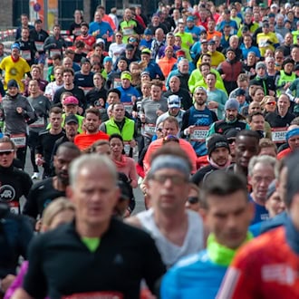 Eine frontale Weitwinkel-Aufnahme einer sehr dichten, großen Menschenmenge, die in bunter Sportbekleidung und mit weißen Startnummern auf der Brust eine asphaltierte Straße entlangläuft. Die Läuferinnen und Läufer tragen Laufshirts, vereinzelt Sportbrillen und Stirnbänder. 