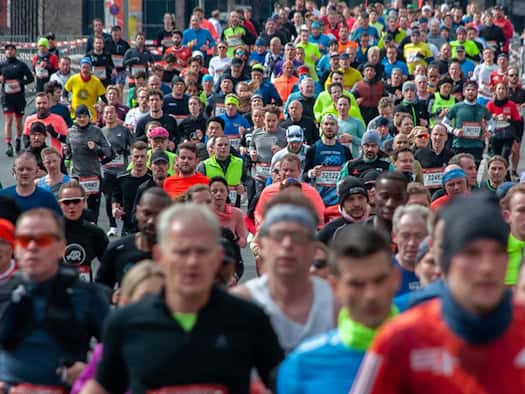 Eine frontale Weitwinkel-Aufnahme einer sehr dichten, großen Menschenmenge, die in bunter Sportbekleidung und mit weißen Startnummern auf der Brust eine asphaltierte Straße entlangläuft. Die Läuferinnen und Läufer tragen Laufshirts, vereinzelt Sportbrillen und Stirnbänder. 