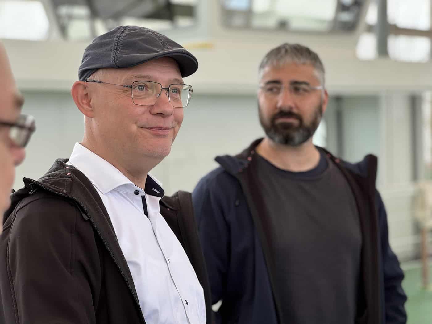 Robert Roch bei der Generalüberholung der Schloßfähre Porträtaufnahme eines lächelnden Mannes mit Schiebermütze und Brille. Im unscharfen Hintergrund steht ein weiterer Mann mit Bart in einer Werfthalle.