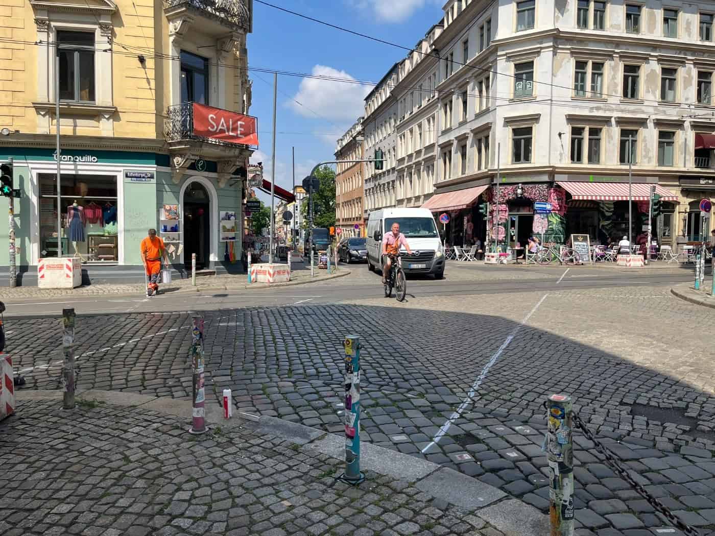 Blick auf eine gepflasterte Straßenkreuzung in Dresden-Neustadt. Ein Arbeiter in orangefarbener Warnkleidung zieht eine weiße Linie auf dem Boden, während im Hintergrund ein Fahrradfahrer und ein weißer Transporter fahren.
