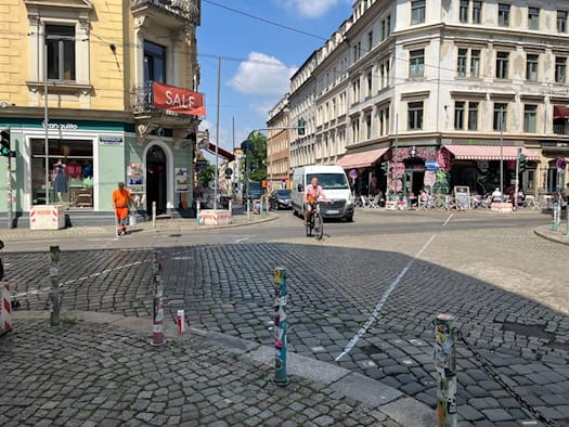 Blick auf eine gepflasterte Straßenkreuzung in Dresden-Neustadt. Ein Arbeiter in orangefarbener Warnkleidung zieht eine weiße Linie auf dem Boden, während im Hintergrund ein Fahrradfahrer und ein weißer Transporter fahren.