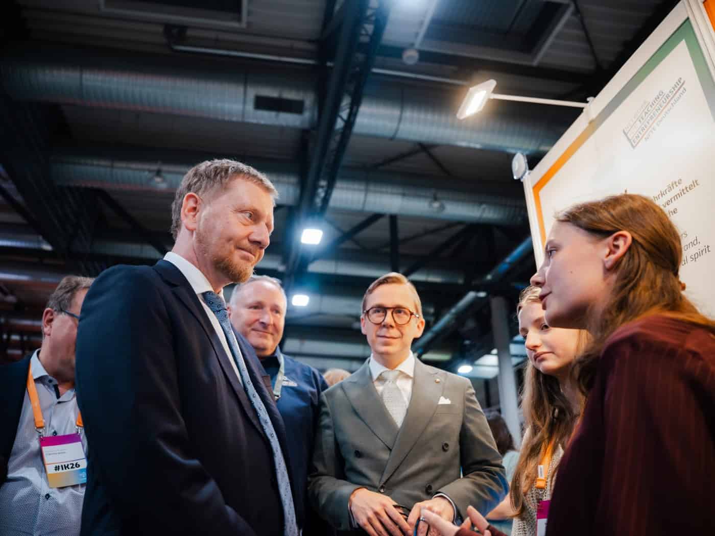 Ministerpräsident Michael Kretschmer (links) und Philipp Amthor im grauen Anzug mit Brille (Mitte) im Gespräch mit einer jungen Frau an einem Messestand in einer großen Halle.