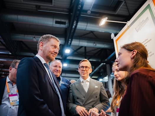 Ministerpräsident Michael Kretschmer (links) und Philipp Amthor im grauen Anzug mit Brille (Mitte) im Gespräch mit einer jungen Frau an einem Messestand in einer großen Halle.