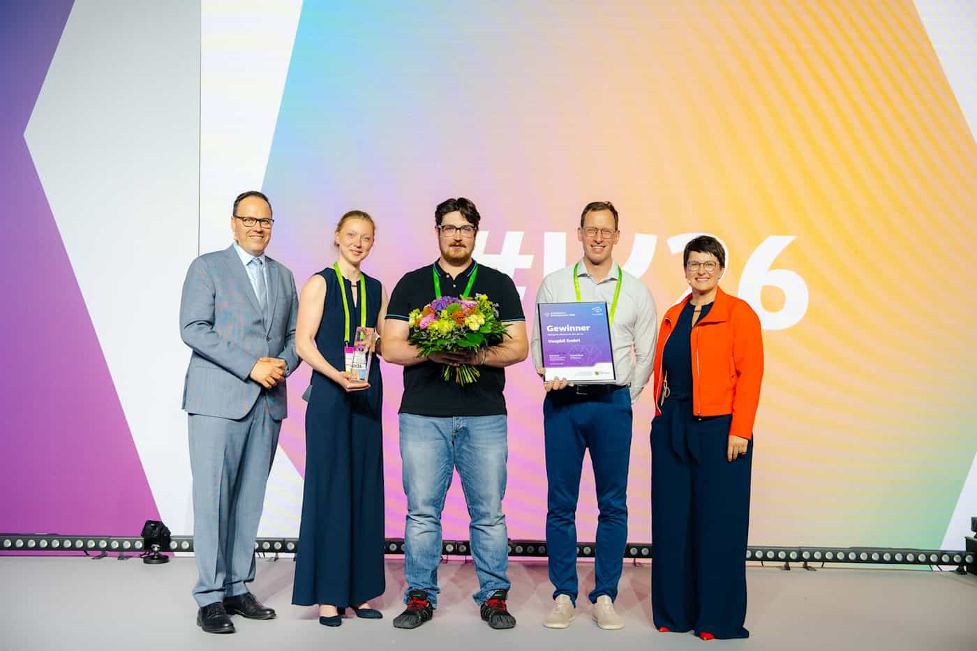 Die Oxaphil GmbH wird als Newcomer des Jahres 2026 ausgezeichnet Eine Gruppe von fünf Personen steht auf einer Bühne. Ein Mann im schwarzen T-Shirt hält einen Blumenstrauß, der man rechts neben ihm präsentiert die Gewinner-Urkunde für die Oxaphil GmbH.