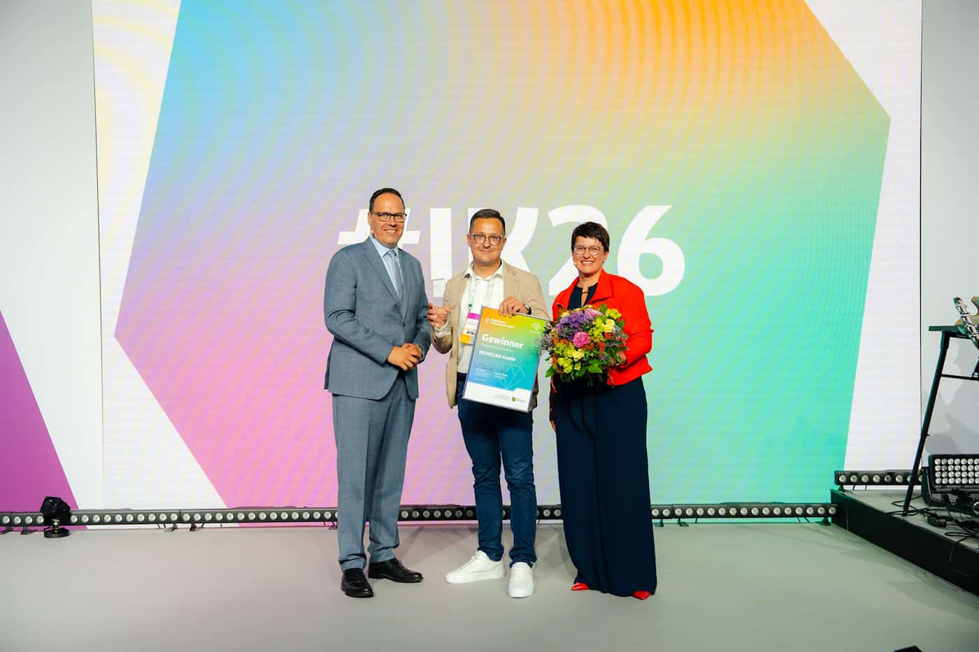 Verleihung des Staatspreises für Gründen 2026 an die DEMECAN GmbH Drei Personen stehen auf einer Bühne vor einer bunten LED-Wand. Der Mann in der Mitte hält stolz eine Gewinner-Urkunde für die DEMECAN GmbH in den Händen, rechts steht eine Frau mit einem Blumenstrauß.