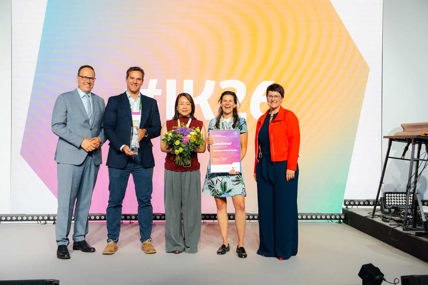 Das Team der Meliodays Medical GmbH erhält den Gründerpreis 2026 Fünf Personen posieren auf einer Bühne. Eine Frau in der Mitte hält einen großen bunten Blumenstrauß, die Frau rechts daneben hält lachend eine Urkunde der Meliodays Medical GmbH hoch.